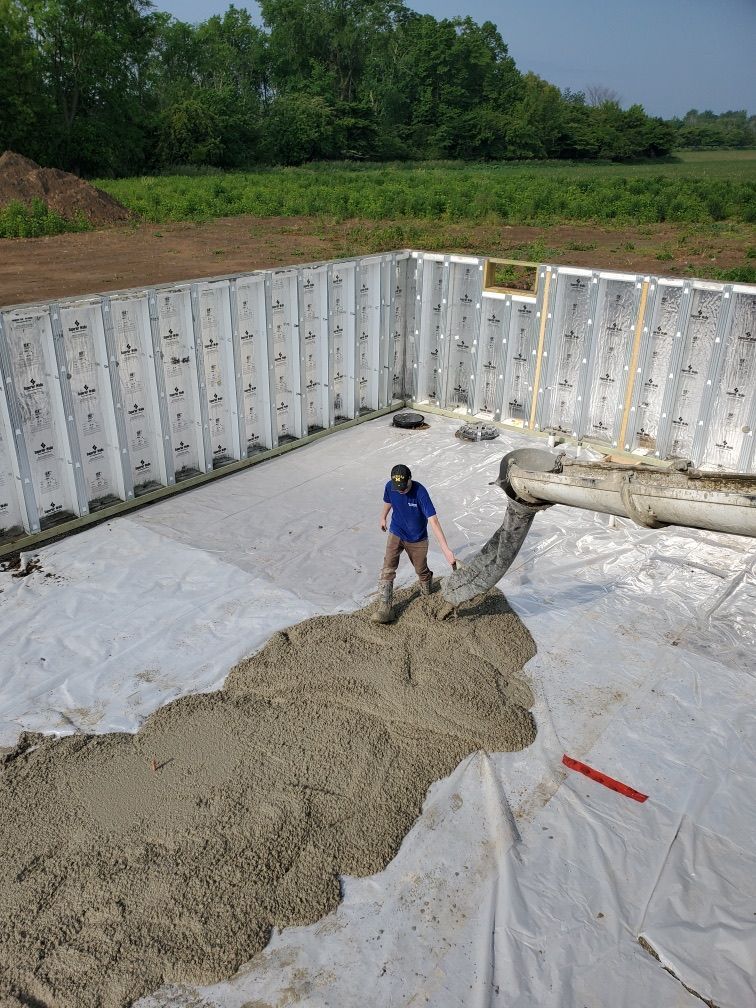 Worker in blue stands in a concrete foundation pit with white retaining forms and a pump hose.