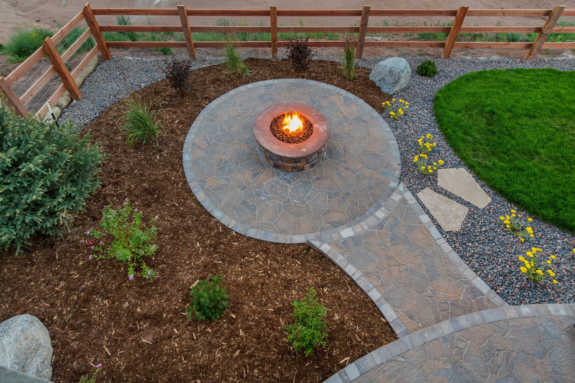 Backyard patio with a lit fire pit, stone walkway, mulch beds, and a wooden fence