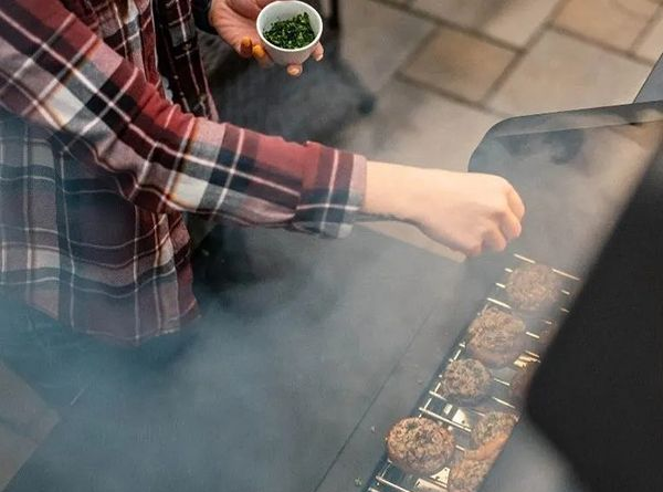 Person grilling skewers over smoky charcoal, wearing a red plaid shirt.