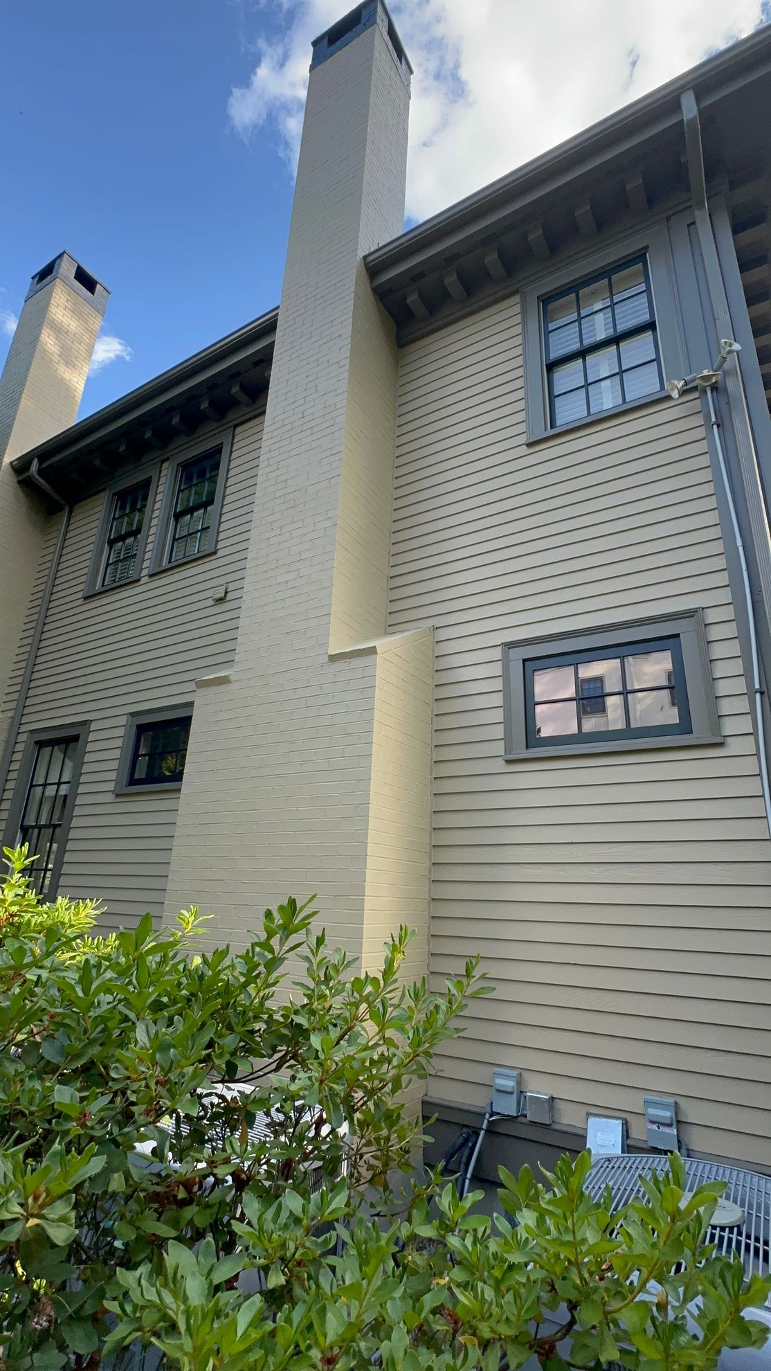 Exterior view of a beige house with a tall chimney against a blue sky, foliage in foreground.