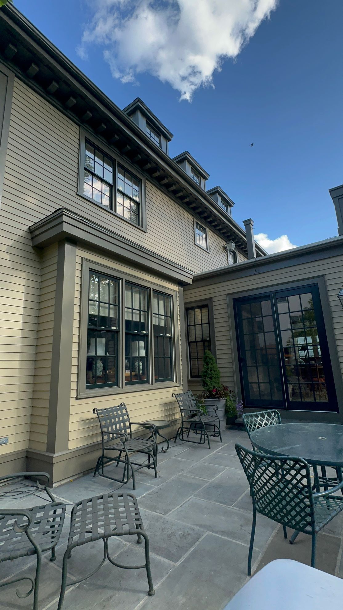 Exterior view of a beige building with a patio, dark trim, and metal chairs under a blue sky with clouds.