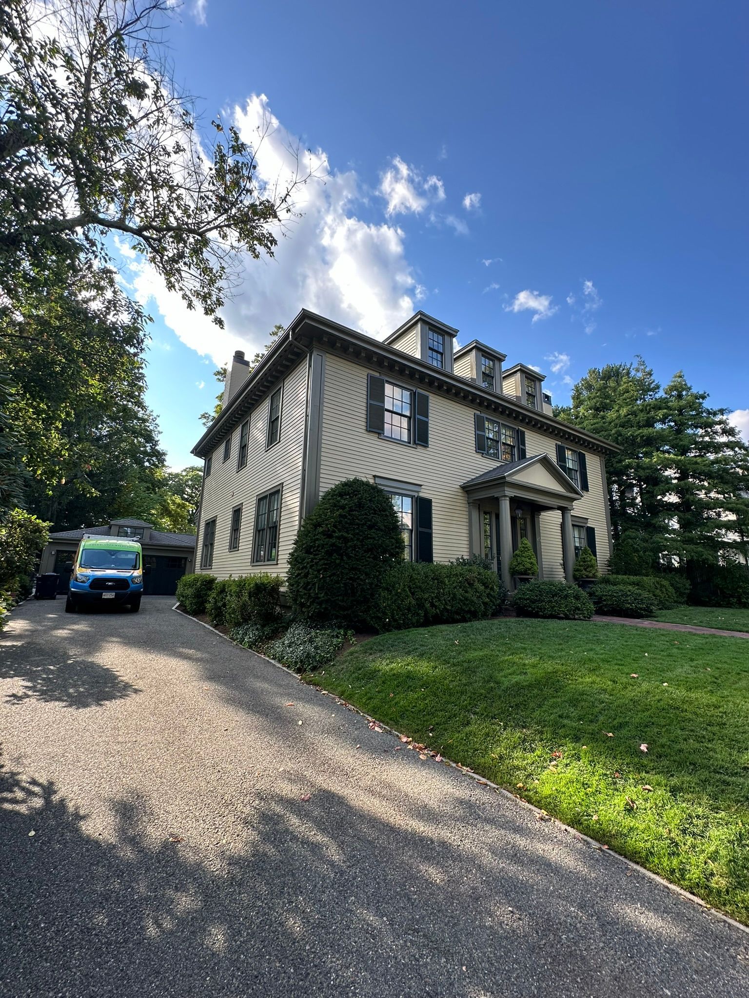 Large two-story house with columns, a gravel driveway, and a well-manicured lawn under a sunny, blue sky.