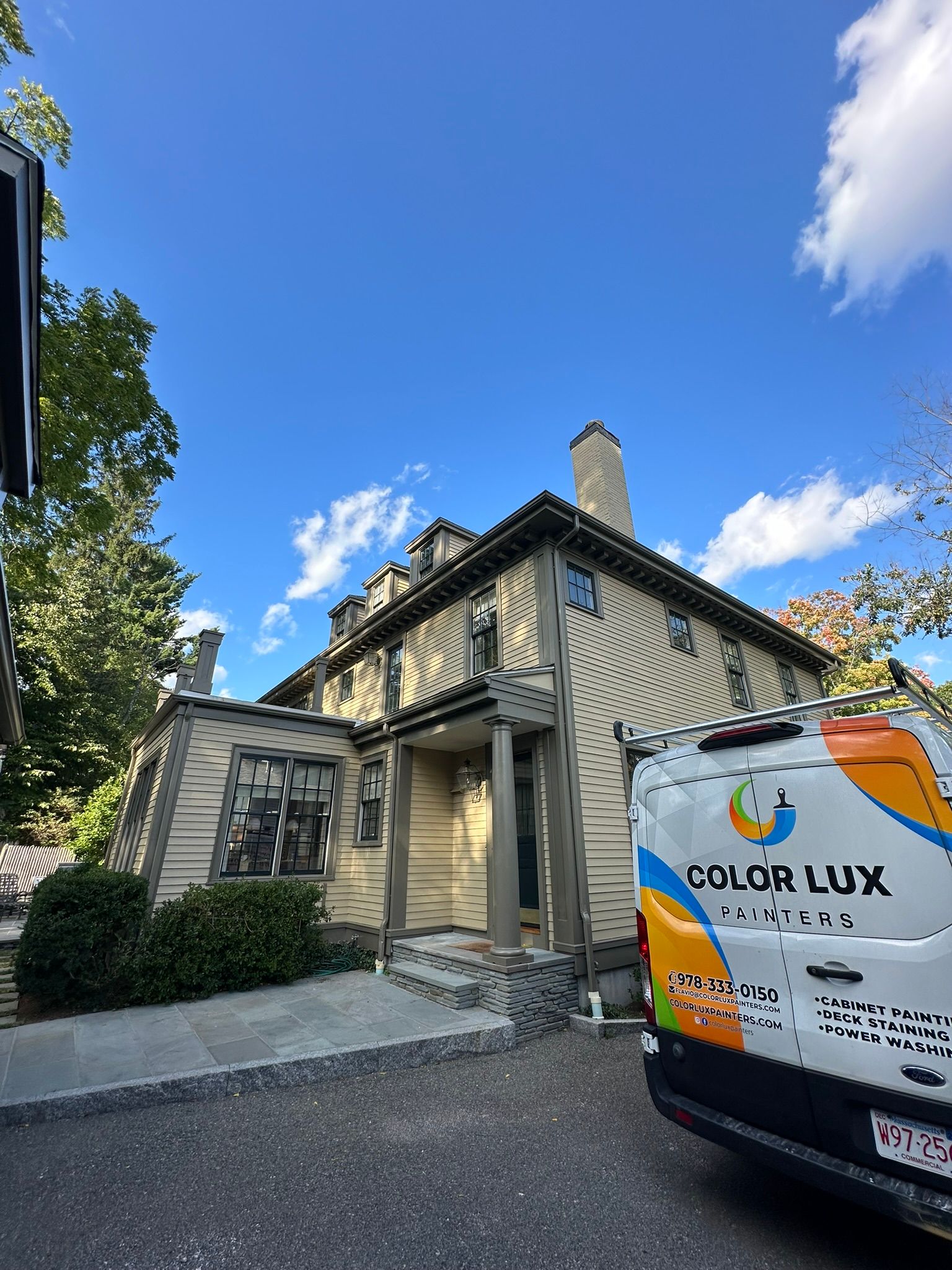 Two-story beige house with gray trim, a Color Lux van parked in front, under a blue sky.