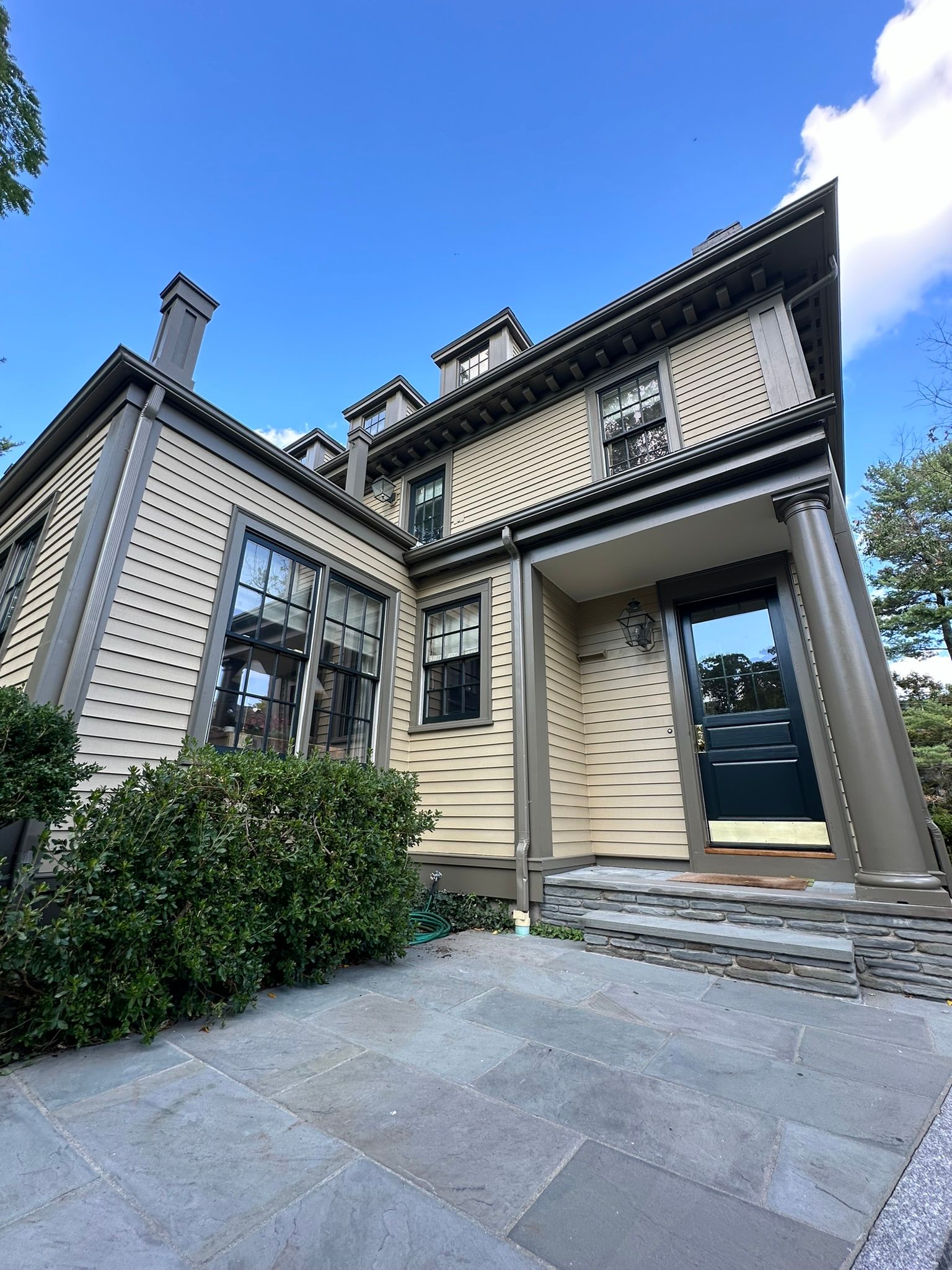 Two-story beige house with dark trim and front porch, set against a blue sky, stone walkway, and green bushes.