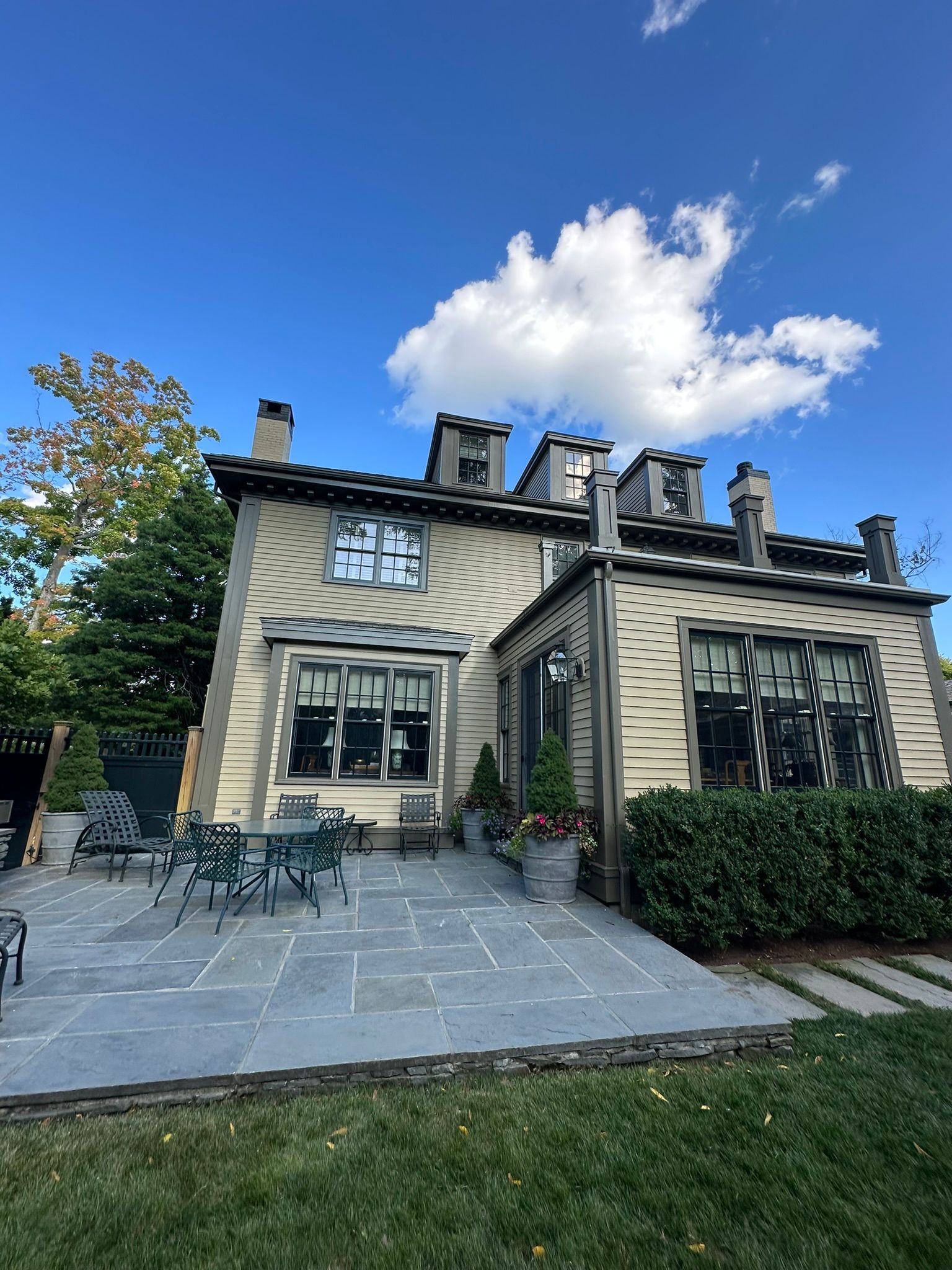 Two-story house with light-colored siding, several windows, and a patio with outdoor seating on a sunny day.