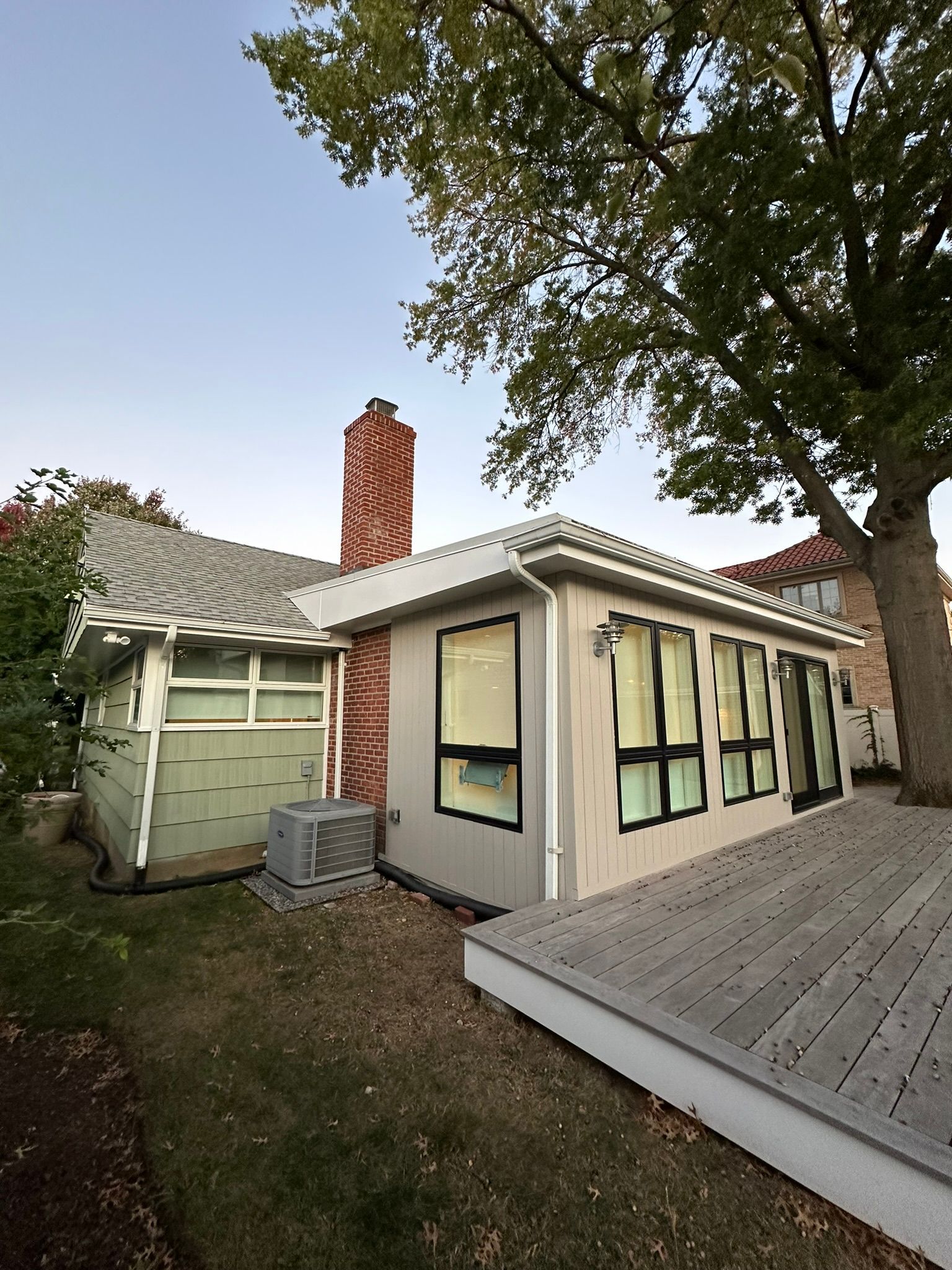 Exterior view of a house with a deck. A light-colored, newly built addition with black-framed windows is on the right. A brick chimney rises above.