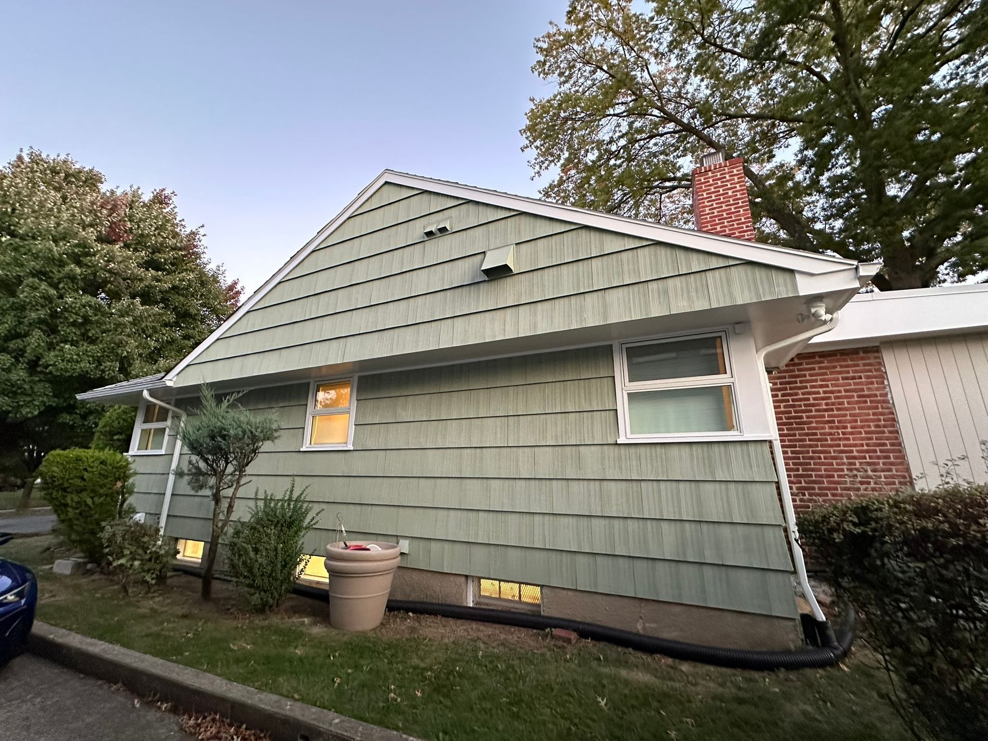 Green-sided house with a red brick chimney and white trim, set in a yard with trees and bushes.
