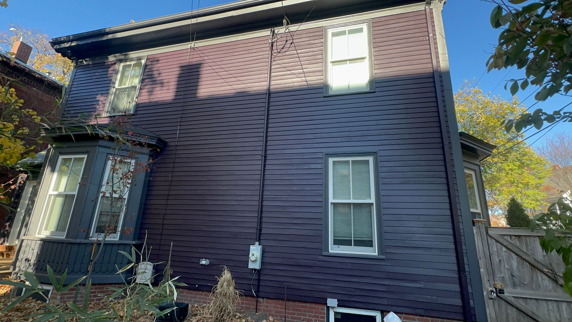Dark purple house exterior with white-framed windows, surrounded by trees and a wooden fence on a sunny day.