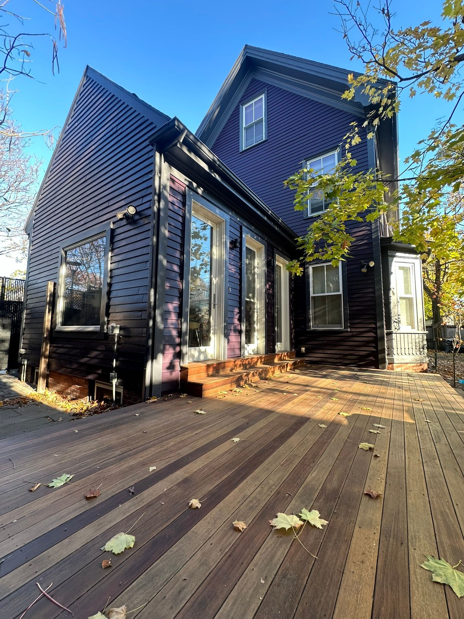 Black house with angled roof, large windows, and a wooden deck scattered with leaves.