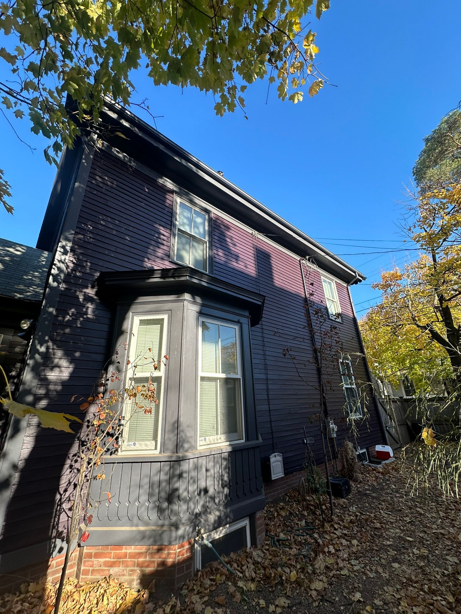 Black house with bay window, set among trees with fall foliage. Sunny day.