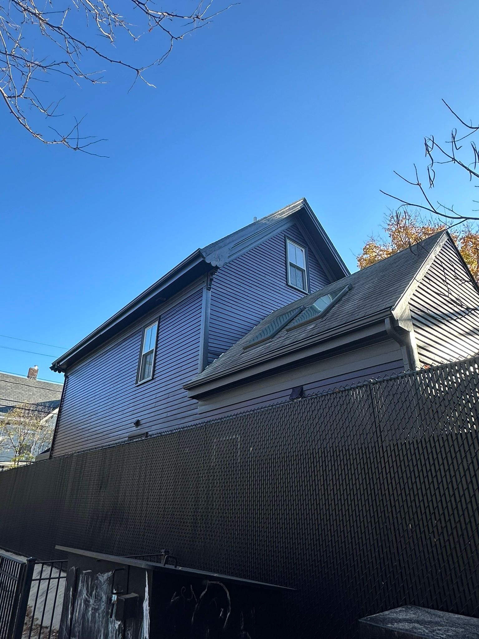 Dark-colored house with two stories and a small shed in front. Blue sky and tree branches overhead.