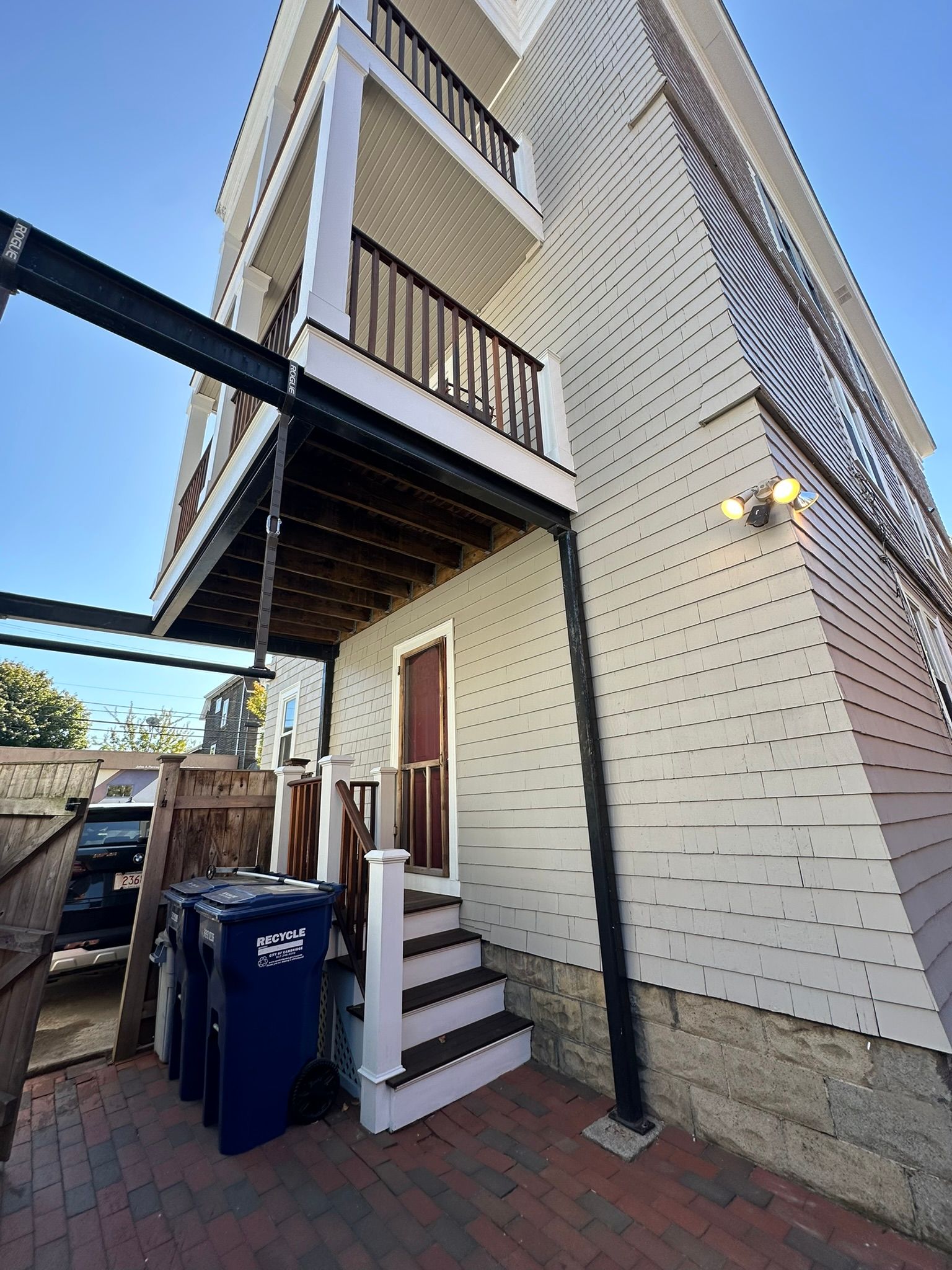 Exterior view of a three-story building with balconies, stairs leading to a door, and trash cans.