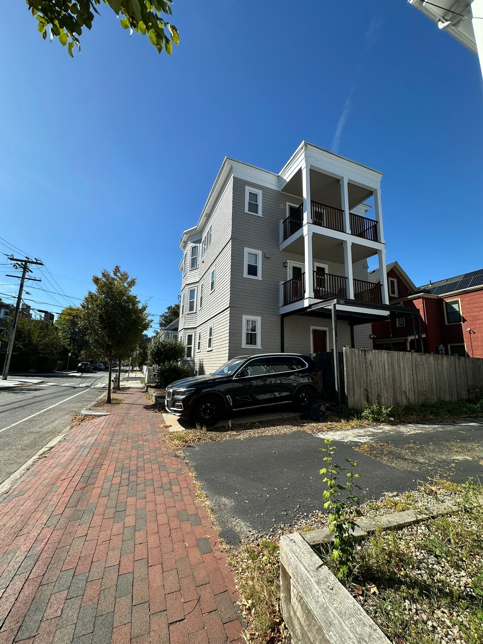 Three-story beige building with balconies, parked cars, and a brick sidewalk on a sunny day.