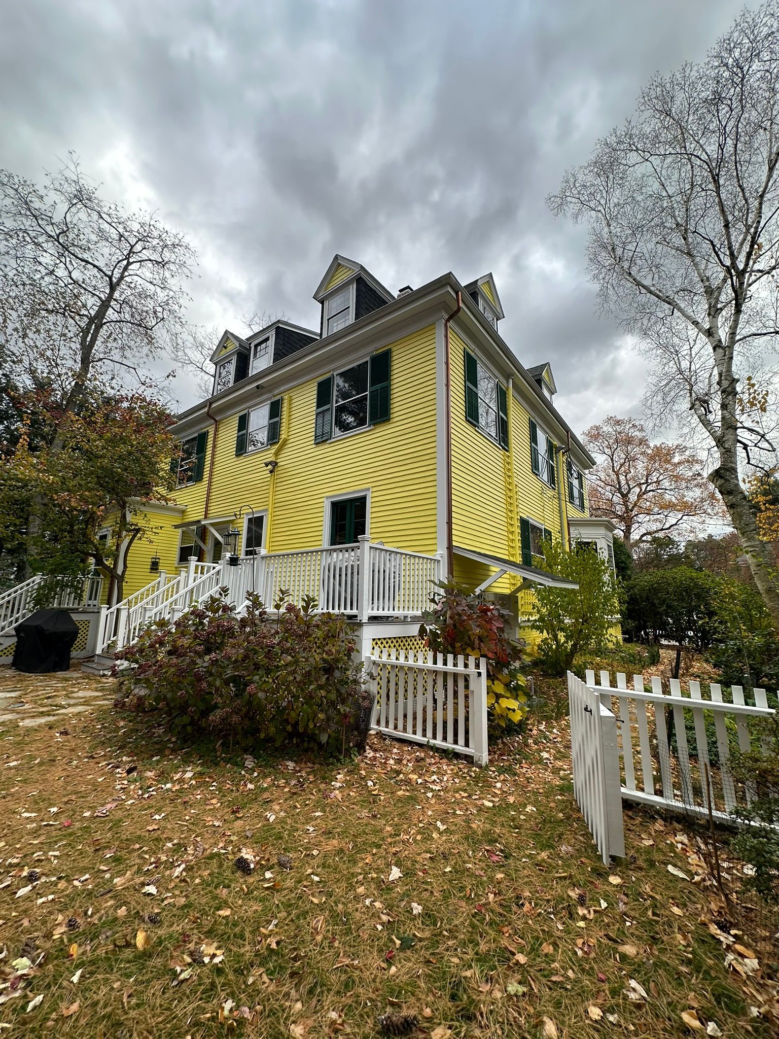 Yellow house with white trim and picket fence under cloudy sky. Leaves on the ground.
