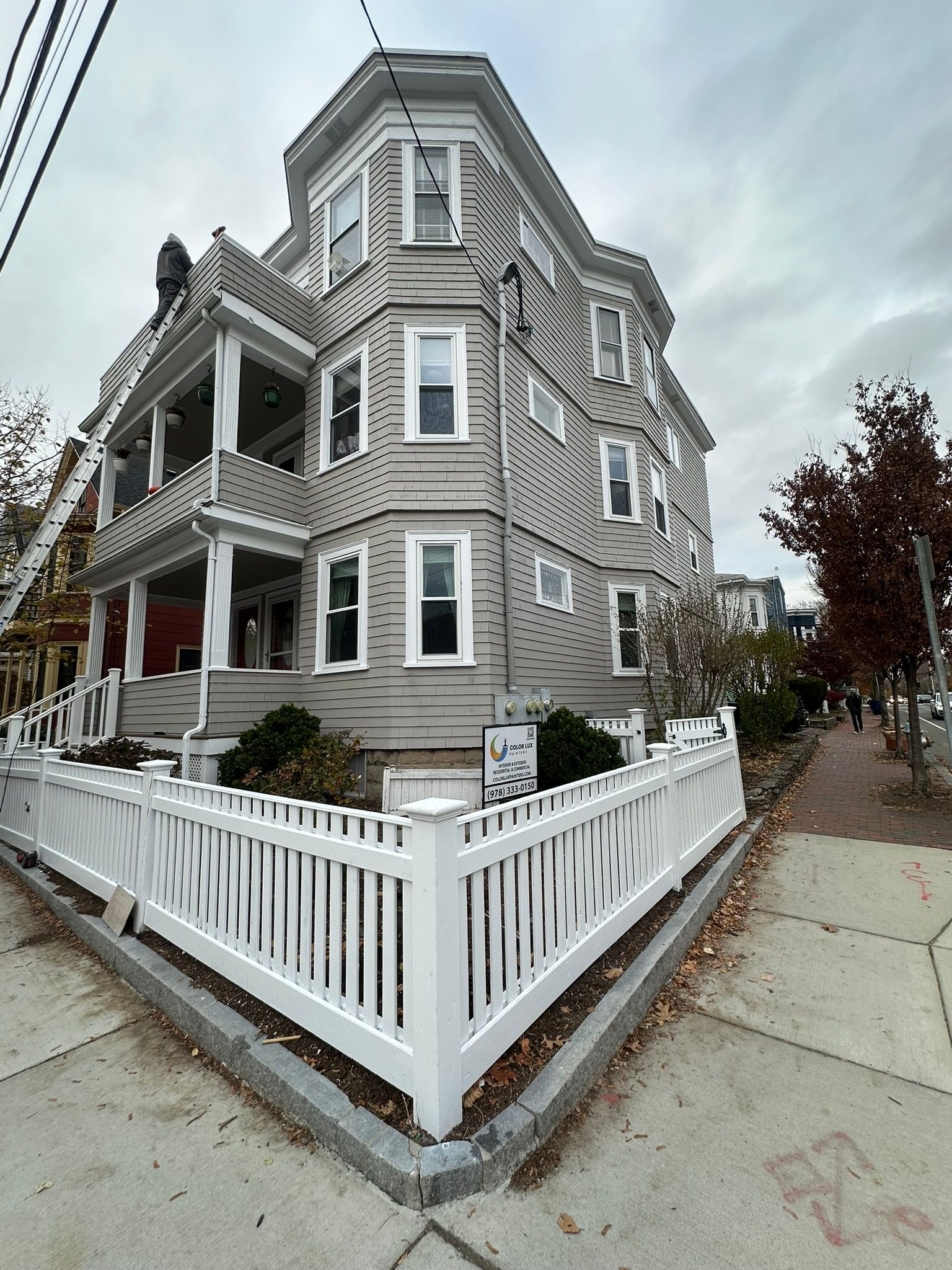 Multi-story grey building with white trim, porch, and picket fence on a corner lot under overcast skies.