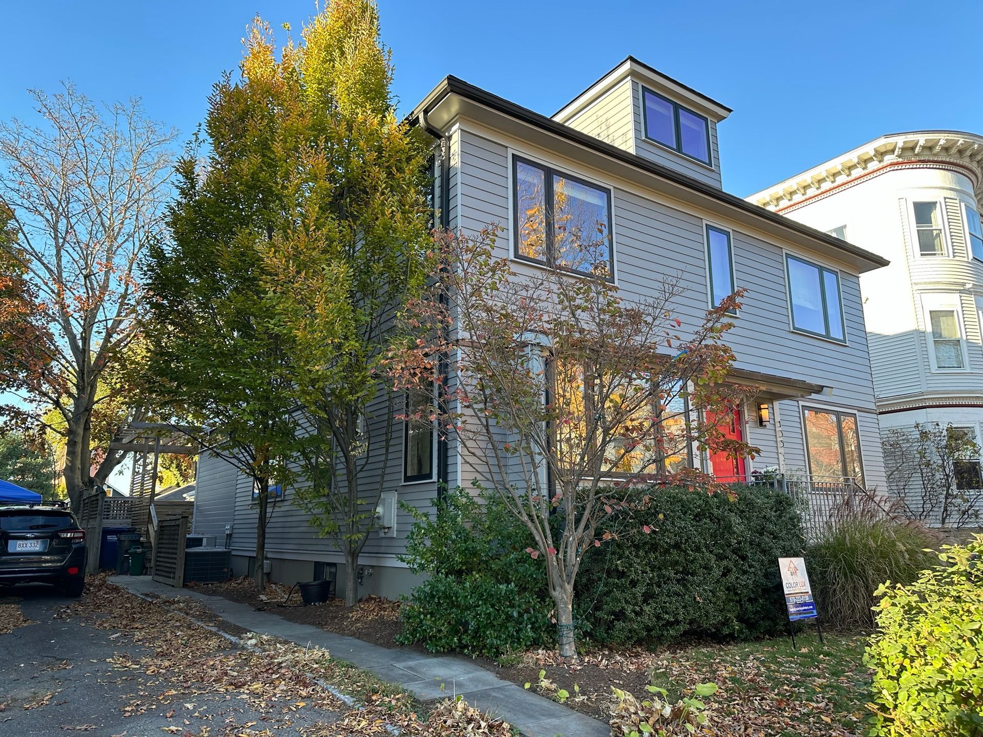 Two-story house with gray siding, red door, and a small tower. Trees and foliage in front.