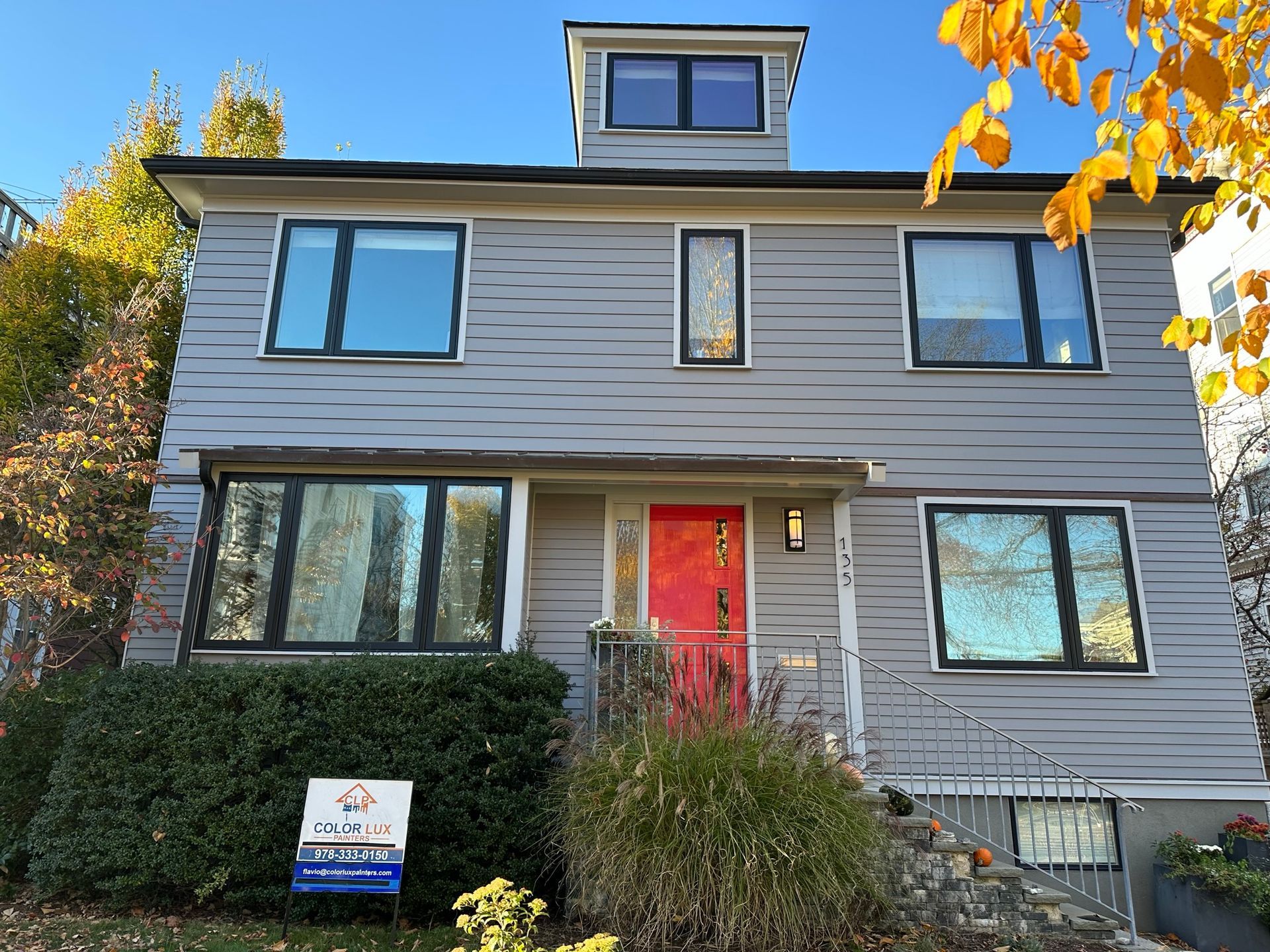 Two-story gray house with red door, black windows. Green bushes in front with yellow fall leaves.