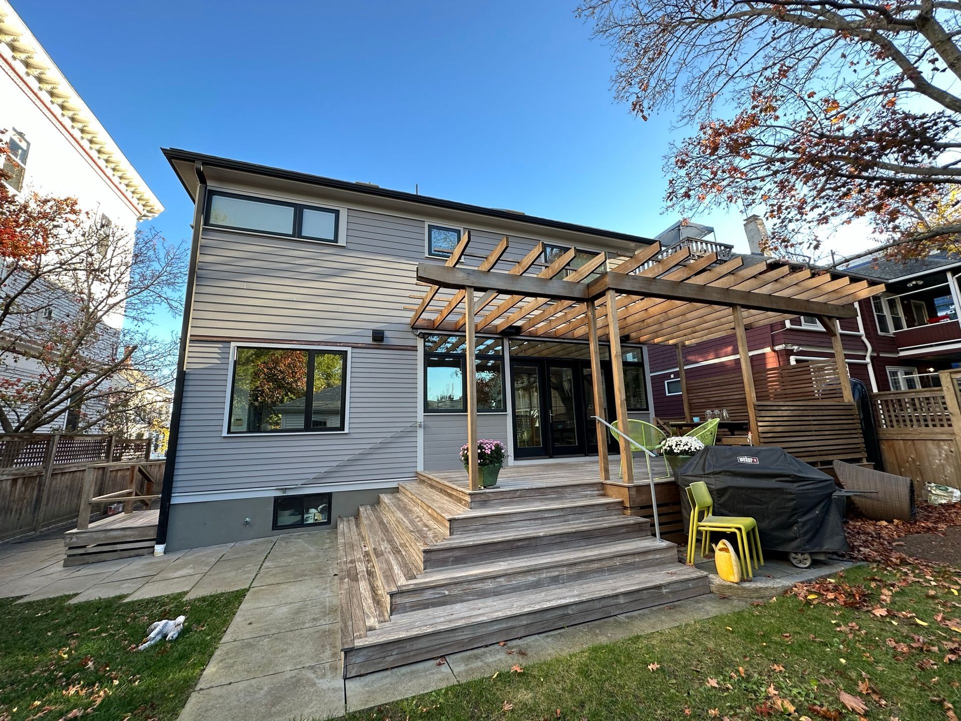 Back of a house with gray siding, wooden deck, pergola, and outdoor grill on a sunny day.
