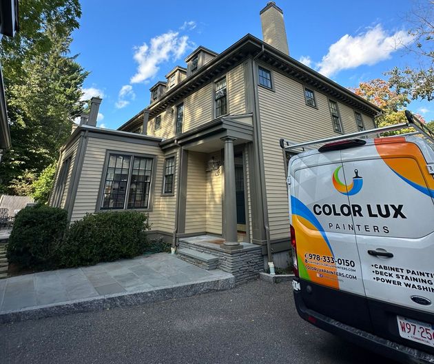 House being painted by Color Lux Painters van, light beige siding with gray trim, under a blue sky.