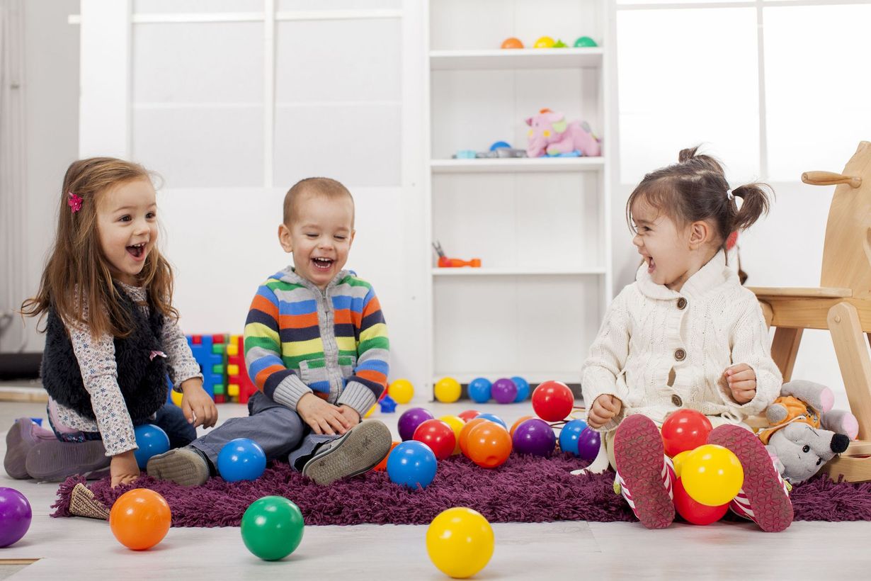 Three young children laughing while playing with colorful balls in a playroom.