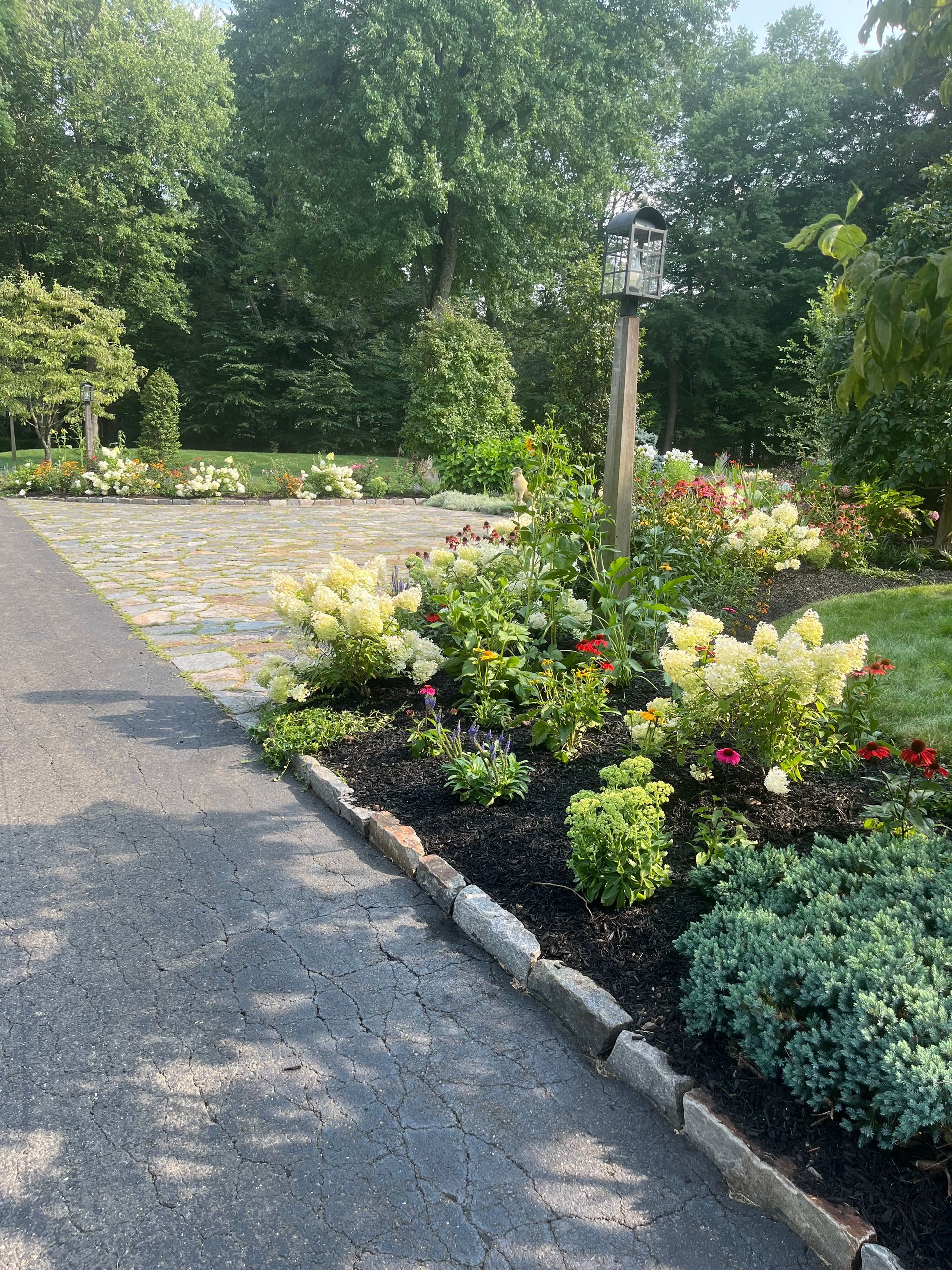 A driveway surrounded by flowers and trees with a birdhouse in the middle.