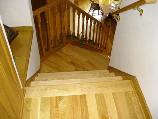 Wooden staircase with light-colored flooring, balusters, and handrail, leading downwards.