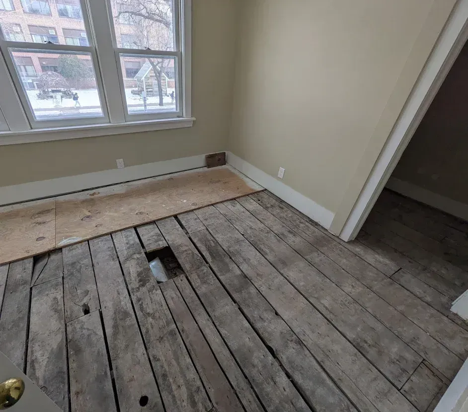 Room with exposed wooden floorboards, a missing section, and an open doorway.  Window with outside view.