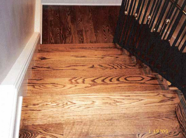 Wooden stairs and landing, dark wood with visible grain. Staircase railing on the right, light wall on the left.