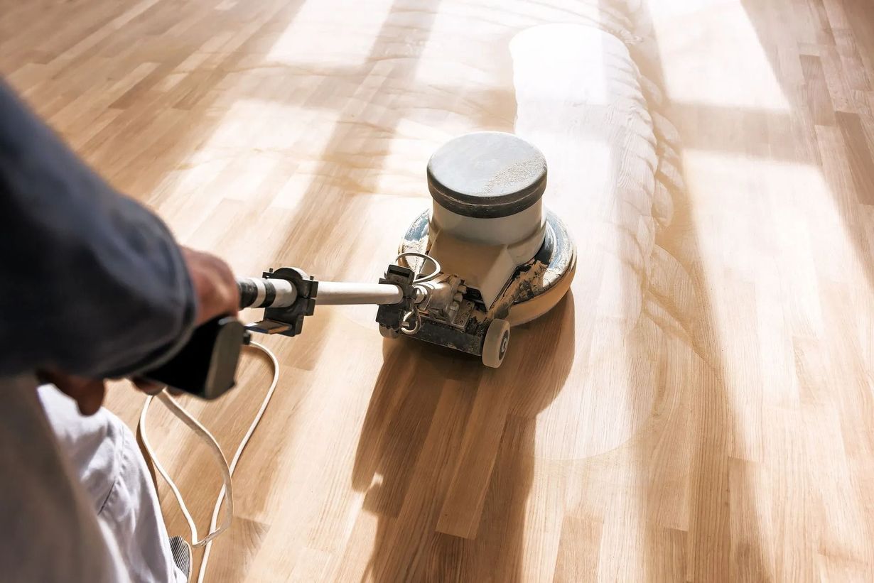 Person using a floor sander on a hardwood floor, sanding in light-filled room.
