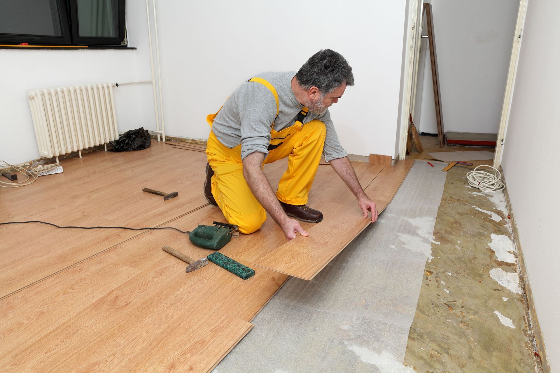 Man installing laminate flooring in a room, with tools and materials visible.