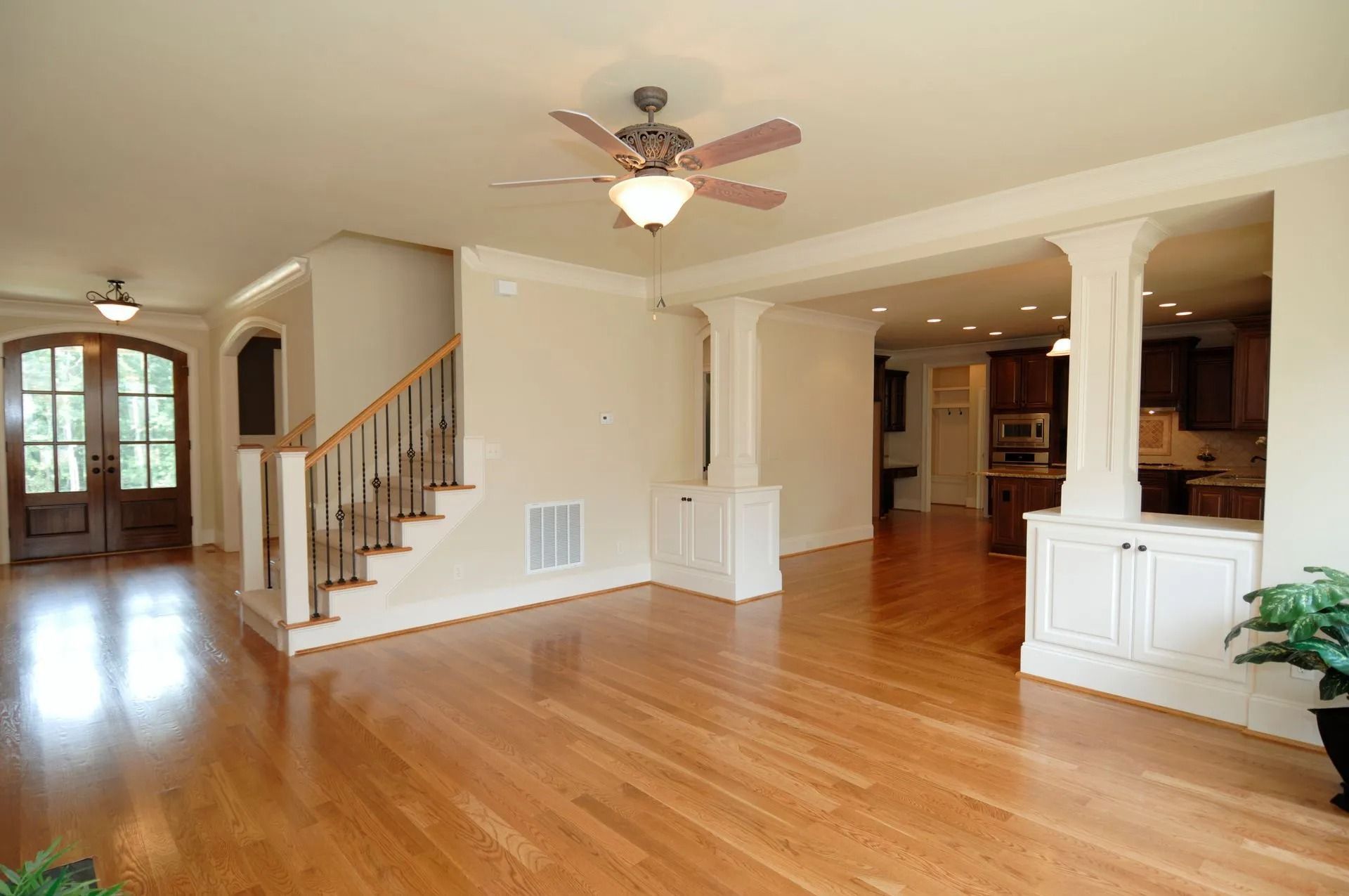 Spacious interior with hardwood floors, stairs, pillars, and a ceiling fan; leading to a kitchen.