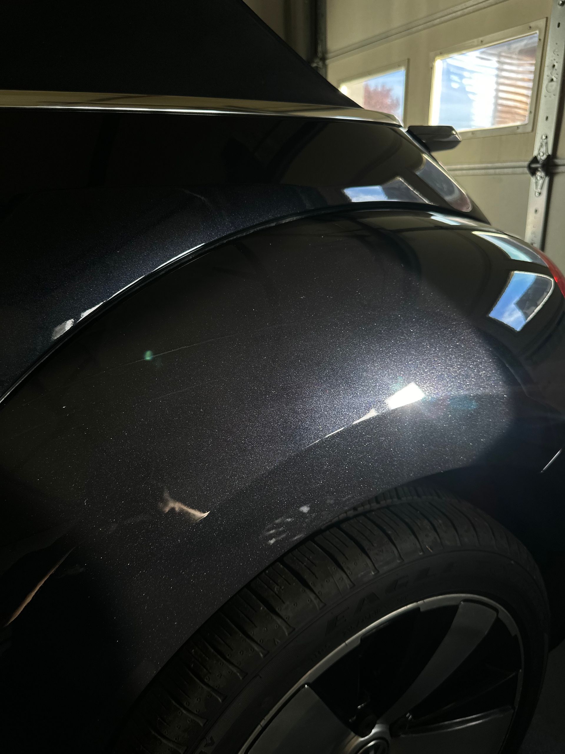 Black car's rear quarter panel, showing tire and shiny paint with light reflecting, indoors.