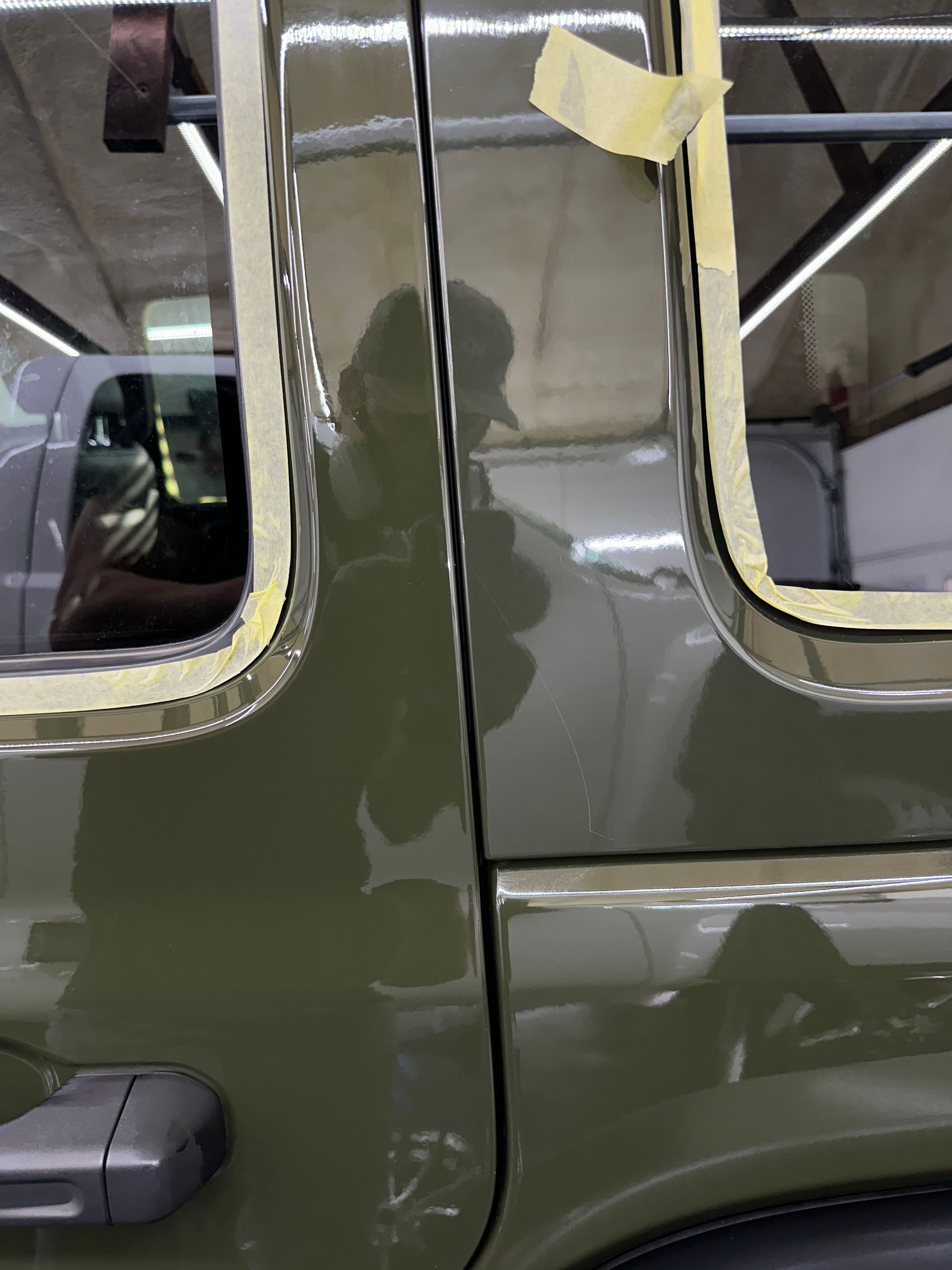 Olive green car door, taped for painting. Reflection of a person visible. Workshop setting.
