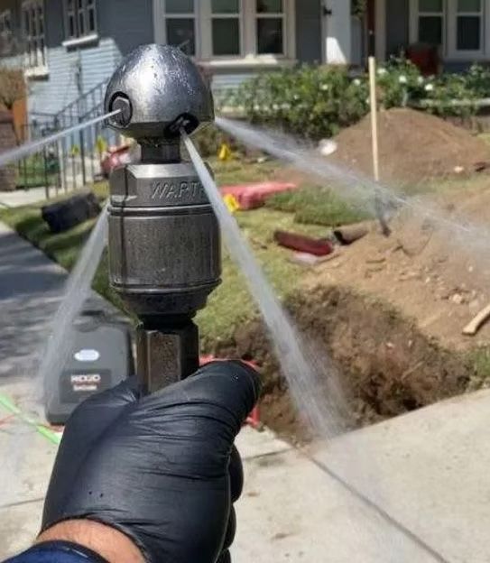 A hand in black gloves holding a nozzle spraying water. The nozzle is pointed towards an open trench.