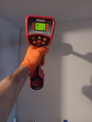 Hand holding a red and black wall scanner; orange glove, white wall.