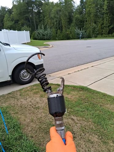 Hand holding a black and silver industrial tool with a coiled hose near a white truck and a driveway.