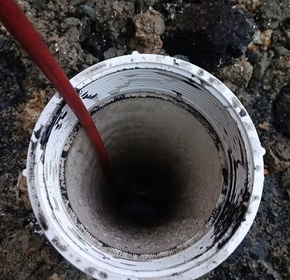 A red tube inserted into a white, threaded pipe set in dirt and rock.