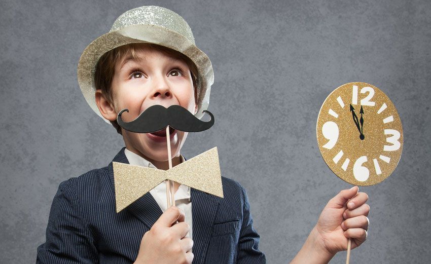 A young boy wearing a fake mustache and bow tie is holding a clock.