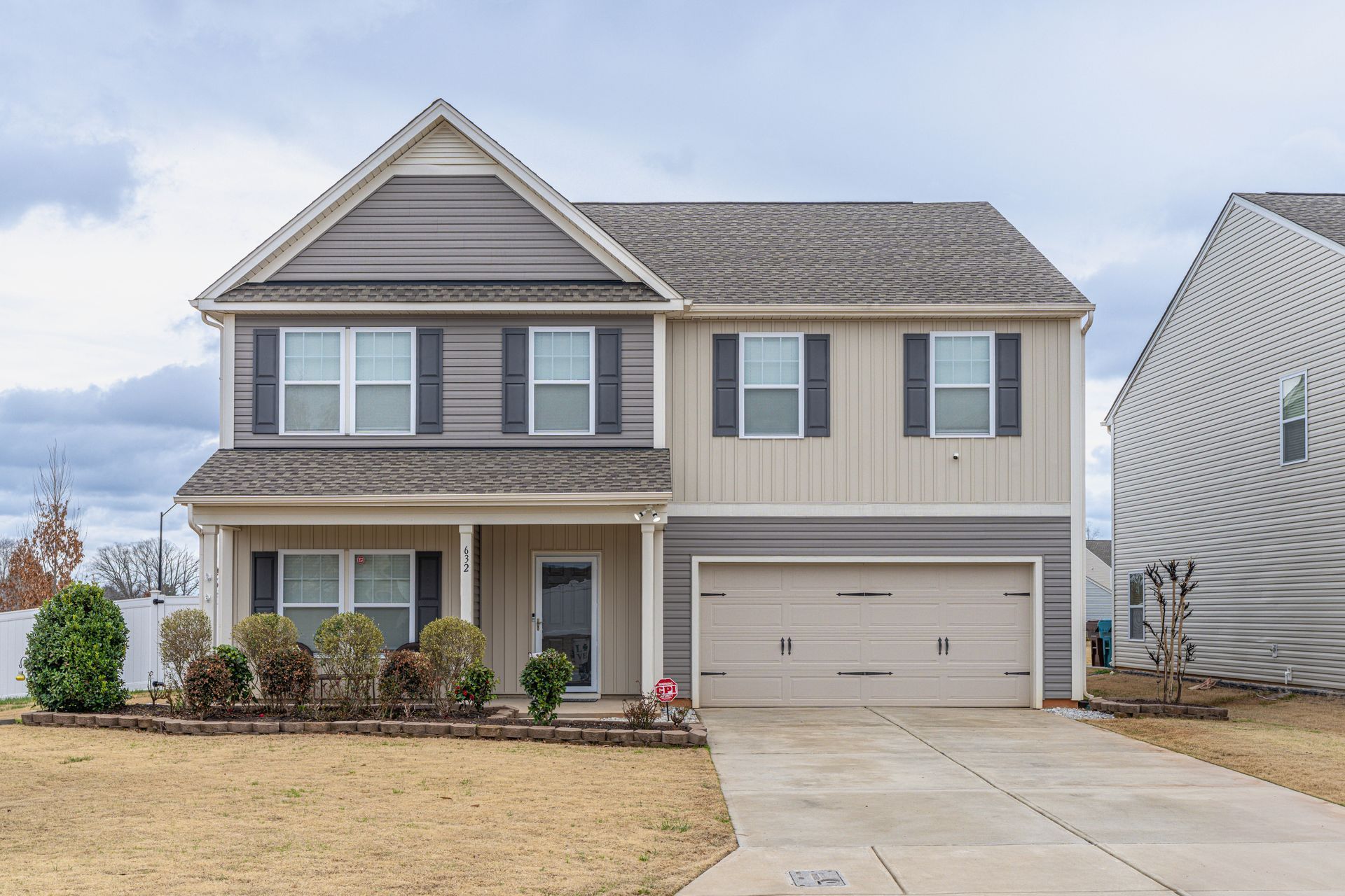 A two-story suburban home with tan and gray siding, a two-car garage, and a landscaped front yard under a cloudy sky.