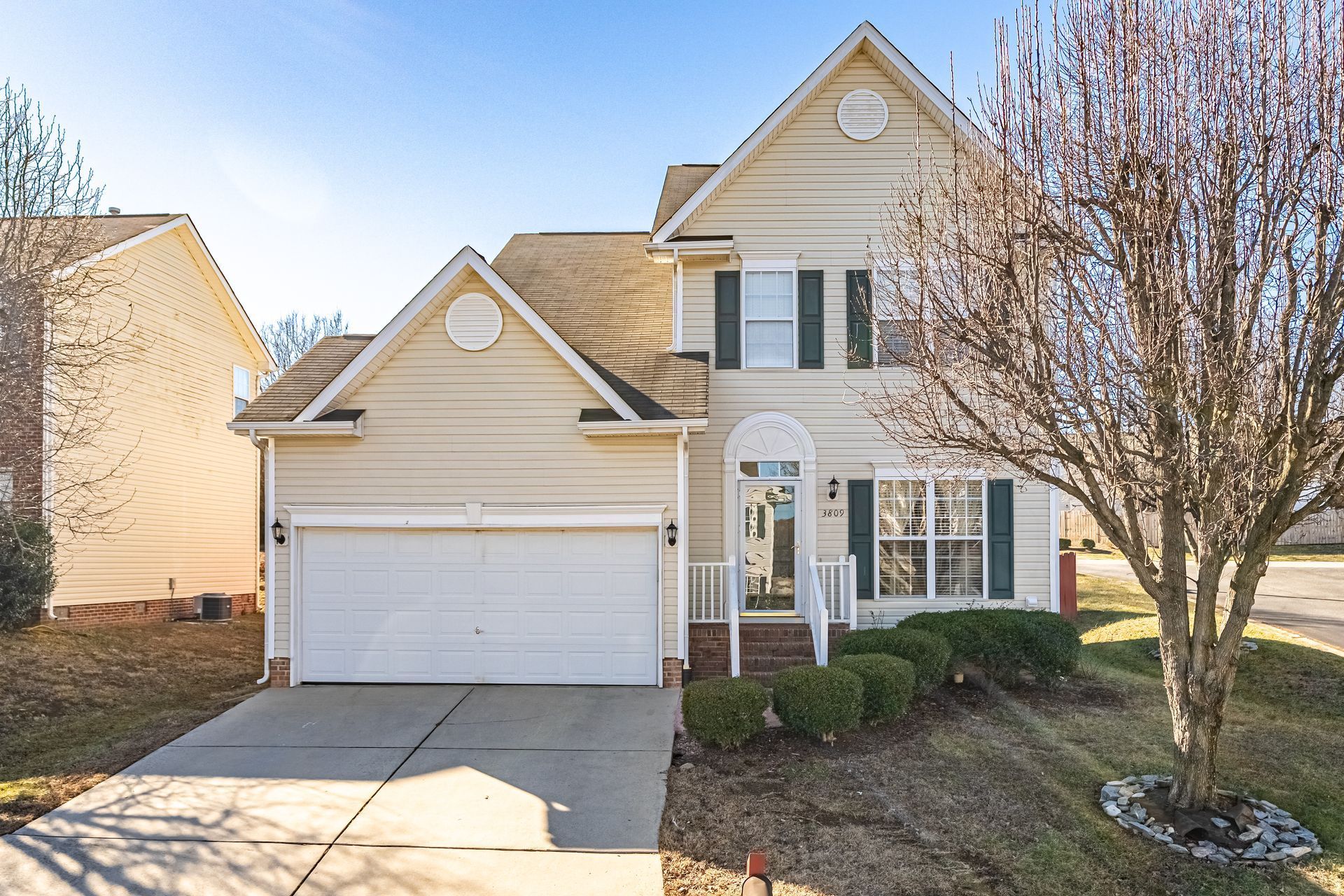 A two-story, light yellow suburban house with a two-car garage, white trim, and a small front yard with a tree.