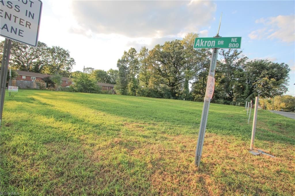 A grassy, vacant lot at the corner of Akron Street, with trees in the background and a traffic sign on the left.