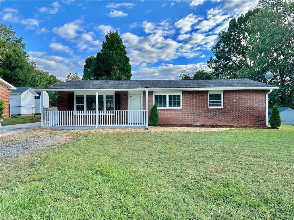 Ranch-style brick house with white porch and fence on a grassy lawn under a blue sky.
