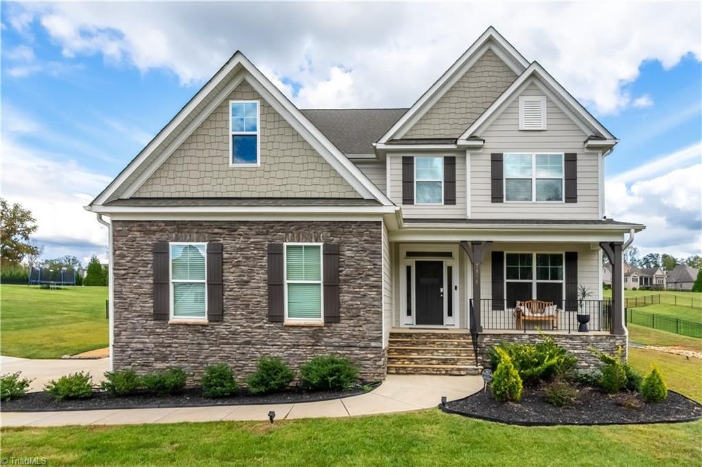 Two-story house with stone and gray siding, black shutters, porch, and landscaped yard under a cloudy sky.