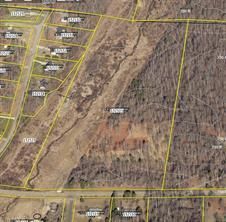 Aerial view of land parcels with houses on the left, a stream through a field, and wooded area on the right