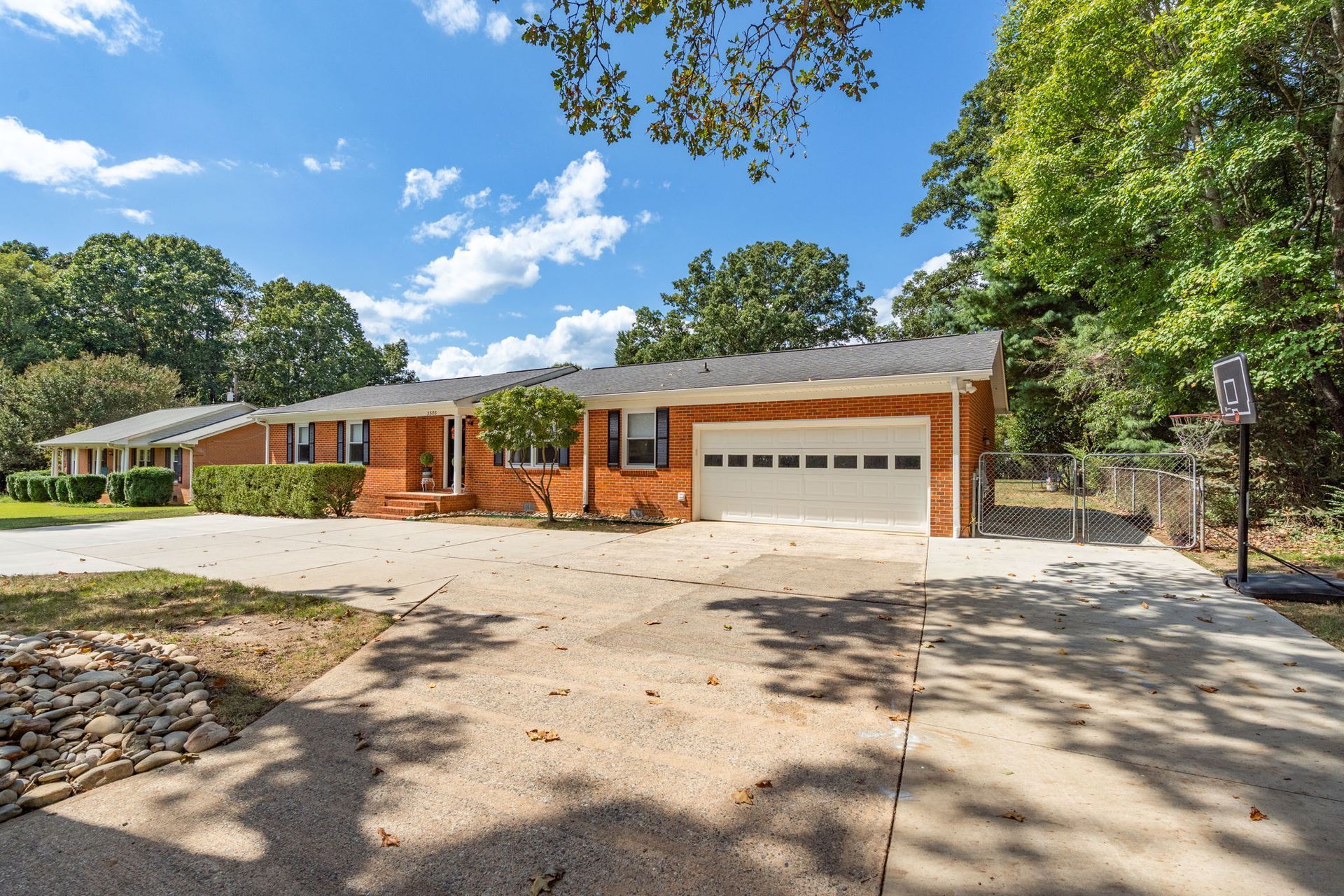 Brick ranch-style house with attached garage, concrete driveway, and surrounding trees under a blue sky.