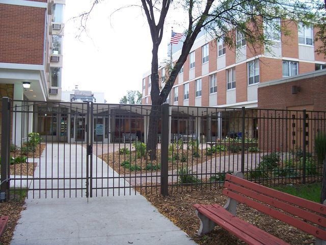 A red bench sits in front of a fence in front of a building