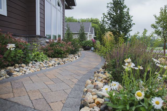 A brick walkway leading to a house surrounded by flowers and rocks.