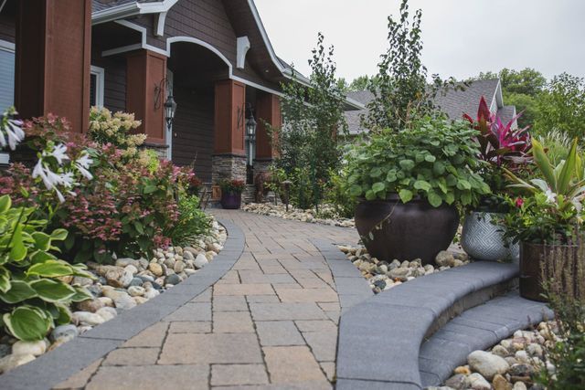 A brick walkway leading to a house surrounded by potted plants and rocks.
