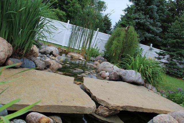 A pond surrounded by rocks and plants with a white fence in the background.