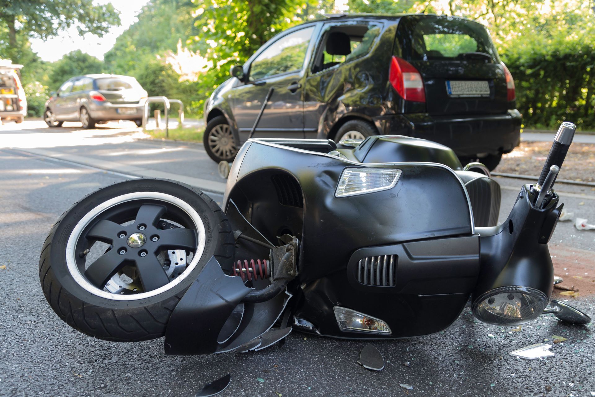 Overturned black scooter in the road with a detached wheel and a black car nearby